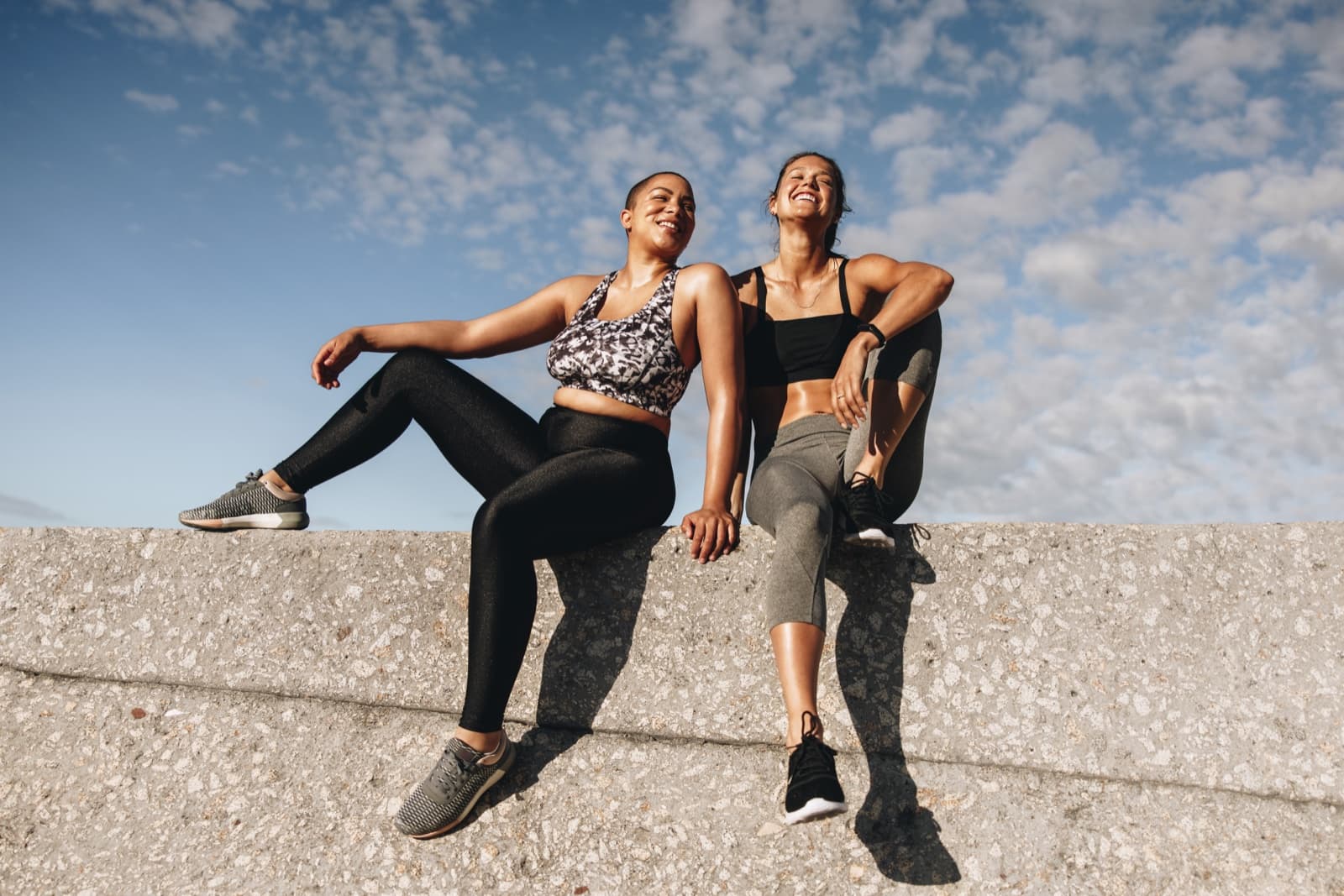 Two women laughing while sitting on a concrete wall in workout clothes