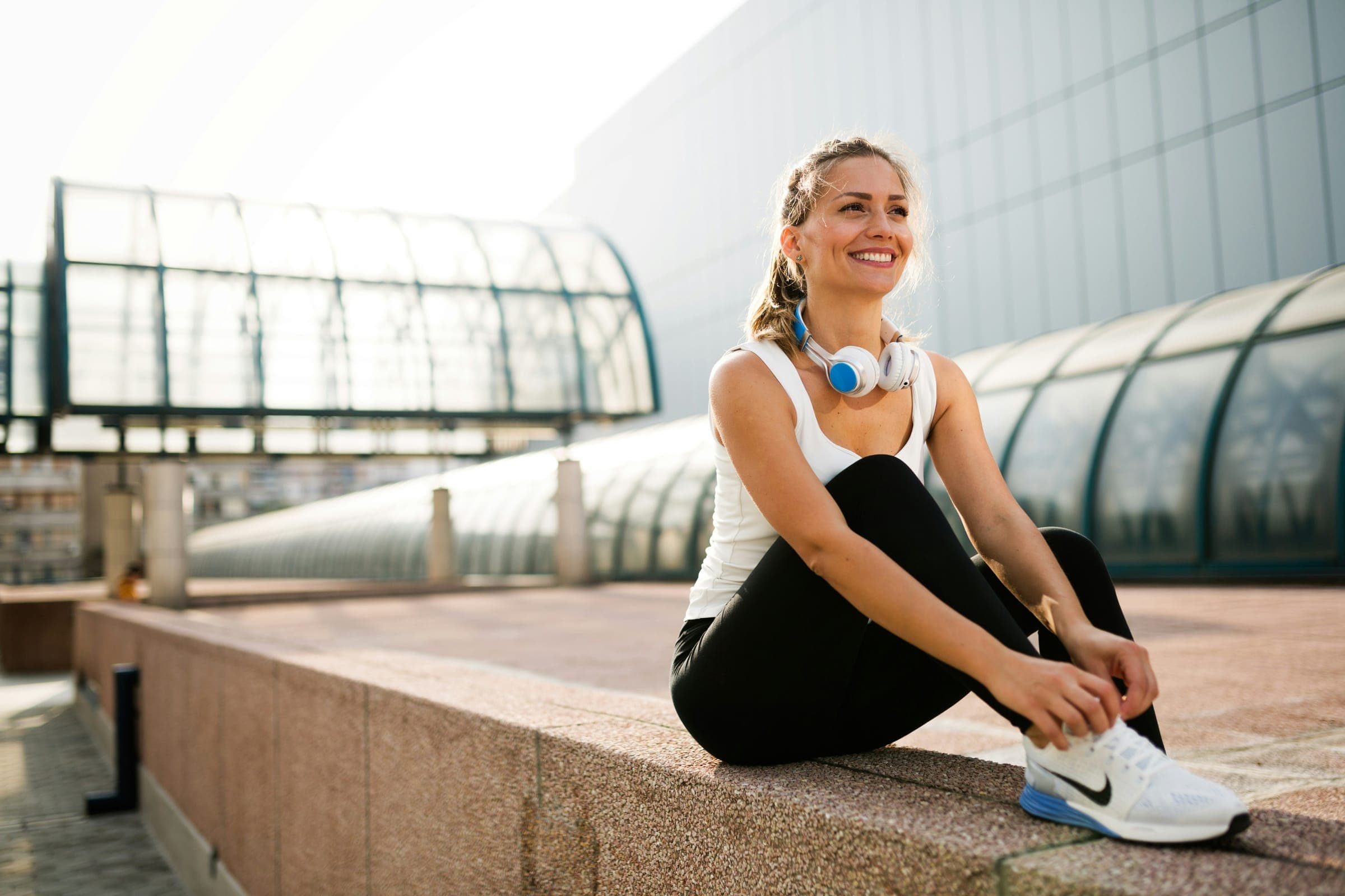 Woman smiling while tying her shoe outdoors, headphones around her neck