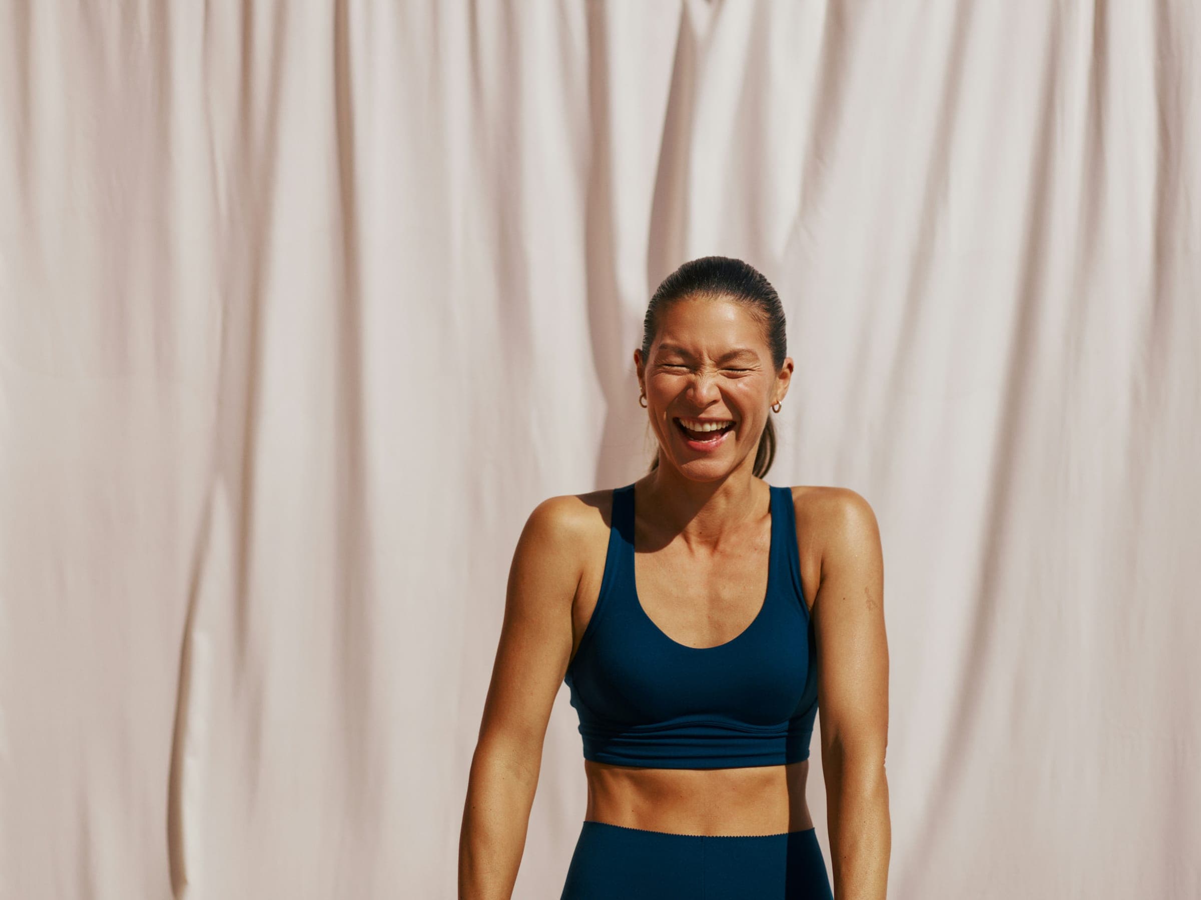 Woman in a navy sports bra laughing in front of a cream curtain
